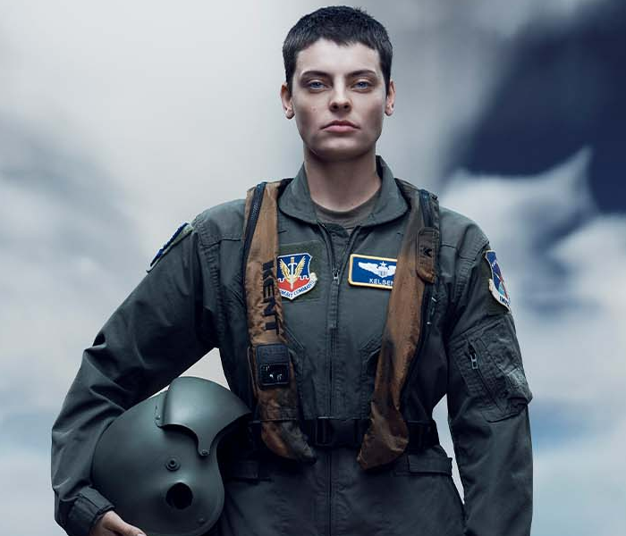 A woman dressed as a fighter pilot looking stoic and standing in front of a cloudy sky