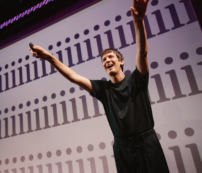 A man in all black with his arms up smiling with a PowerPoint slide of exclamation points behind him