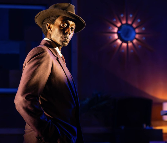 A Black man in a sharp brown suit and hat looking over his shoulder at the camera